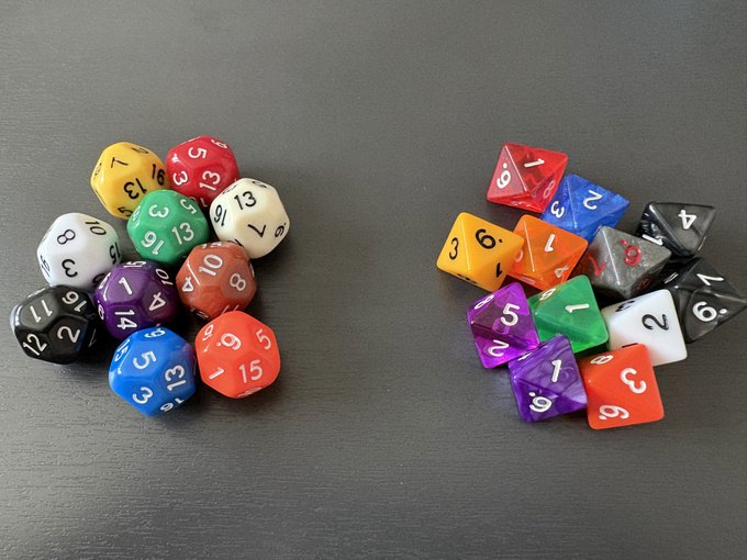 Two piles of polyhedral dice rest on a gray surface, with the left pile featuring larger dice showing higher numbers like 13, 15, and 10, and the right pile containing smaller, more colorful dice displaying numbers like 3, 6, and 2. The dice illustrate the concept that 16-sided dice (16×16) align mathematically with the 2,048 words in the BIP39 wordlist used for Bitcoin seed phrases.