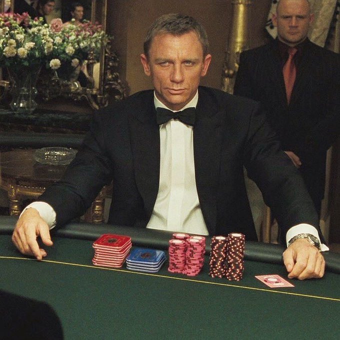 A man in a black tuxedo and bow tie sits at a green felt poker table with stacks of red and blue chips and a card in front of him, while a bald man in a dark suit stands guard behind him. The image ties into the "GM everyone" post by evoking the high-stakes, risk-taking glamour of casino gambling, fitting the bold energy of announcing a bonus hunt on Stake.