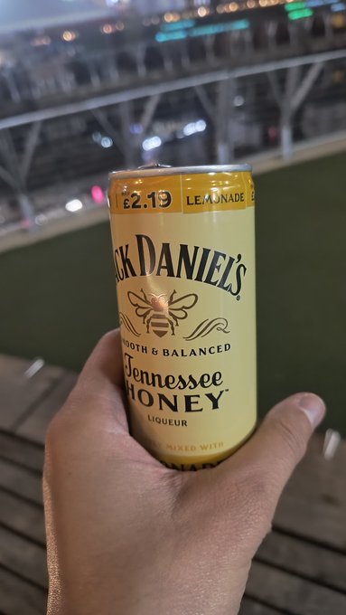 A hand holding a can of Jack Daniel's Tennessee Honey Lemonade priced at £2.19, with a blurred stadium background at night.