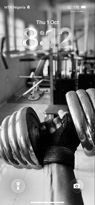 A black and white iPhone lock screen wallpaper displays a photograph of a muscular arm wearing a black glove lifting a heavy dumbbell in a gym, with the time 8:12 and date Thu 1 Oct visible on the MTN Nigeria LTE network.
