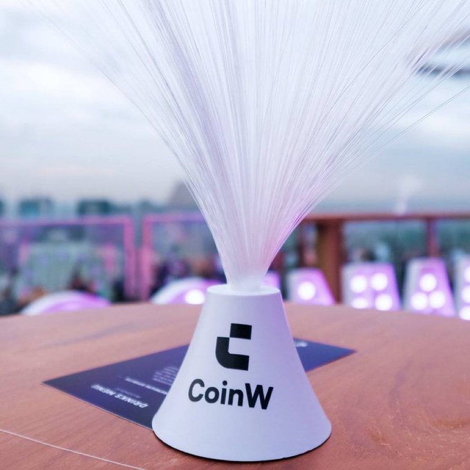 A white cone-shaped decorative lamp with fiber optic strands emitting light, branded with the "CoinW" logo on a wooden table.
