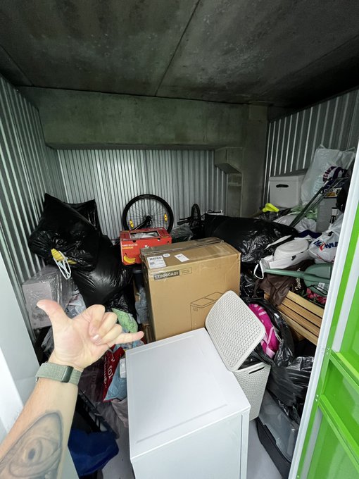 A person's tattooed arm points toward a cluttered storage unit filled with trash bags, boxes, a bicycle wheel, and miscellaneous items.