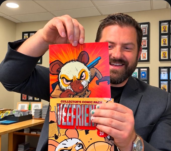 A smiling bearded man in a black blazer holds up a 'VEEFRIENDS' Collector's Comic Pack featuring a cartoon rat character, posing in what appears to be an office with framed collectibles on the wall behind him.