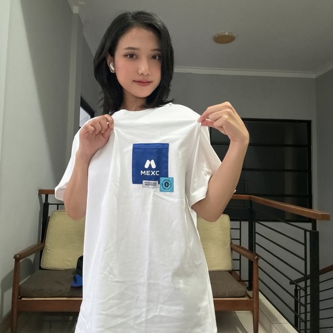 A young woman holds up a white MEXC t-shirt in an indoor setting, appearing to showcase the branded merchandise.