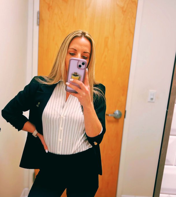 A woman takes a mirror selfie in professional attire—a black blazer over a striped button-up blouse—holding a lavender case phone in front of a wooden door, embodying a confident "GM" (good morning) vibe to start the workday.