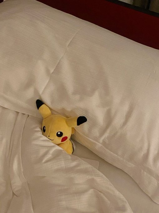 A small plush Pikachu toy peeks out from underneath white bed sheets on a mattress.