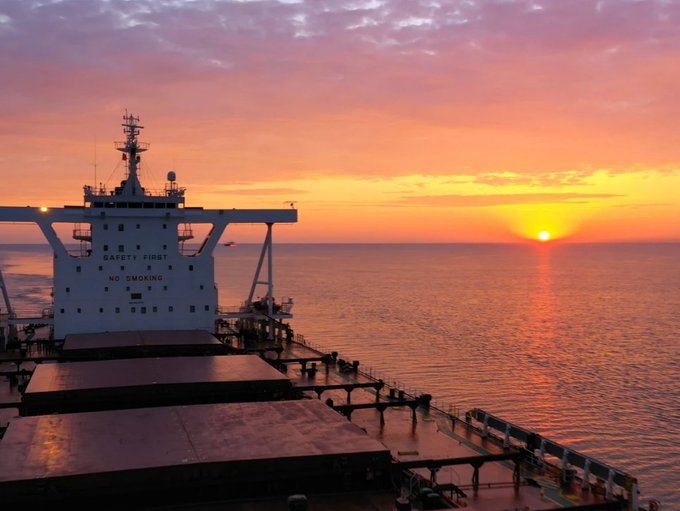 A scenic photograph taken from a cargo ship at sunset, showing the ship's superstructure with 'SAFETY FIRST' and 'NO SMOKING' text, overlooking a vibrant orange and pink ocean horizon.
