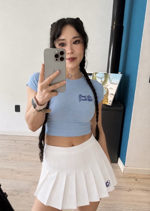 A woman with long braided hair takes a mirror selfie wearing a light blue 'Good Luck Have Fun' cropped t-shirt, a white pleated tennis skirt, and an Apple Watch, holding an iPhone Pro.