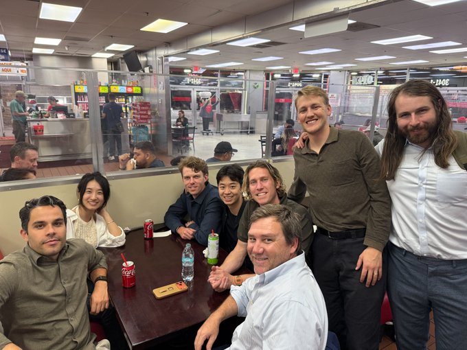 A diverse group of eight people pose smiling around a table at what appears to be Tacos El Gordo, with Coca-Cola cans, water bottles, and a phone visible on the table, while the "Cinn" and "PINK" storefronts in the background suggest a food court setting. The image captures the celebratory post-conference dinner referenced in the caption following Yotta 2026, showcasing the relaxed gathering after a day of deal-making with Hyperbolic Labs.