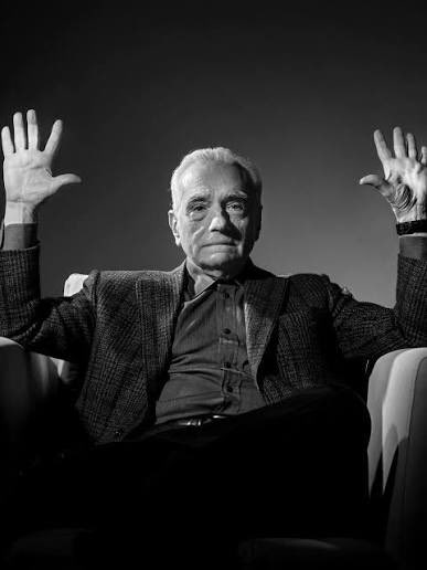 A black-and-white portrait of an older man in a tweed jacket sitting in a chair with both hands raised, palms facing the camera.