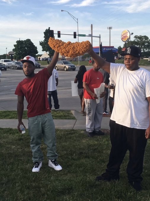 Two young men stand on grass near a street intersection, holding up a massive piece of fried chicken together while onlookers watch in the background near a Burger King sign.