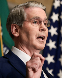 Scott Bessent, the U.S. Treasury Secretary, is shown speaking during what appears to be a press briefing, gesturing with his hand while wearing a dark suit. The image accompanies news of his announcement that the Trump administration plans to seize $1 billion in Iran-linked cryptocurrency this week as part of an effort to isolate and target Iranian financial assets.
