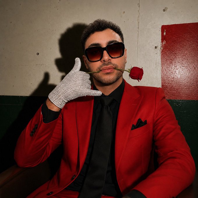 A confident man in a bold red suit and oversized sunglasses poses with a rhinestone glove while playfully holding a rose between his teeth, channeling a flamboyant Valentine's Day aesthetic that humorously contrasts with the post's shy, self-deprecating tone.