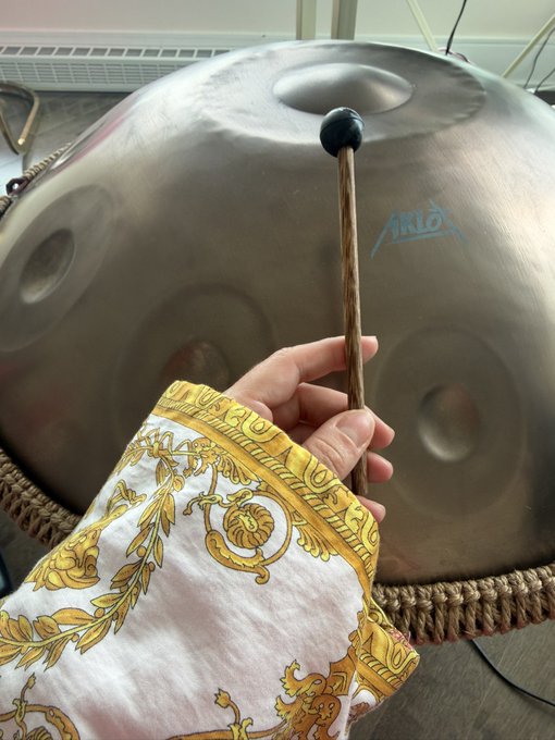 A hand adorned in a gold and white baroque-patterned sleeve holds a wooden mallet with a blue tip poised above an "Arko" brand handpan drum. The image captures a serene moment of musical practice, complementing the post about enjoying a peaceful handpan session while exploring micro coin collecting.