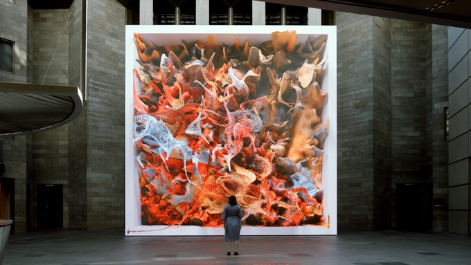 The image shows a woman standing in front of an art installation that appears to be a large screen displaying a digital painting or a 3D rendering of a plant with orange leaves. The artwork seems to be part of a larger display, possibly inside a museum or gallery setting. The woman is looking at the artwork intently, seemingly captivated by it.