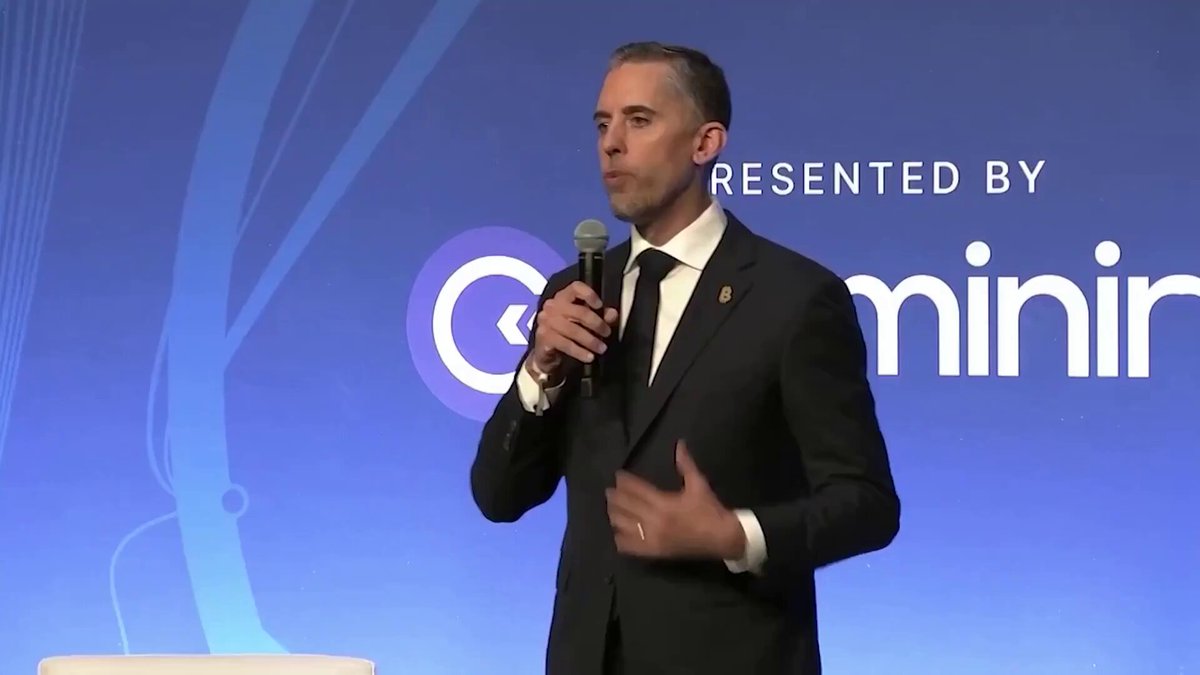 The image features a man dressed in a suit standing behind a podium with his hands at his sides, giving him an authoritative presence as he speaks into a microphone. The background of the image shows a blue wall with white text that reads "BREAKING: $1 BILLION STRIVE JUST BOUGHT OVER 1,100 MORE !!! COUNTING CORPORATIONS ARE BUYING BILLIONS NEW MICHAEL SAYLOR IS HERE". This suggests that the man is addressing an important issue or event related to cryptocurrency and its impact on various companies.