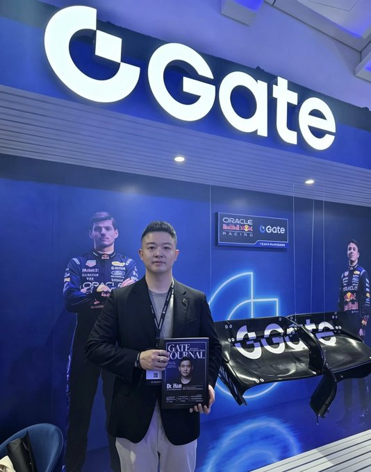 A photograph of an Asian man in a dark blazer holding 'Gate Journal' and a Gate.io branded banner, posing in front of a Gate.io booth with Oracle Red Bull Racing F1 driver imagery in the background.