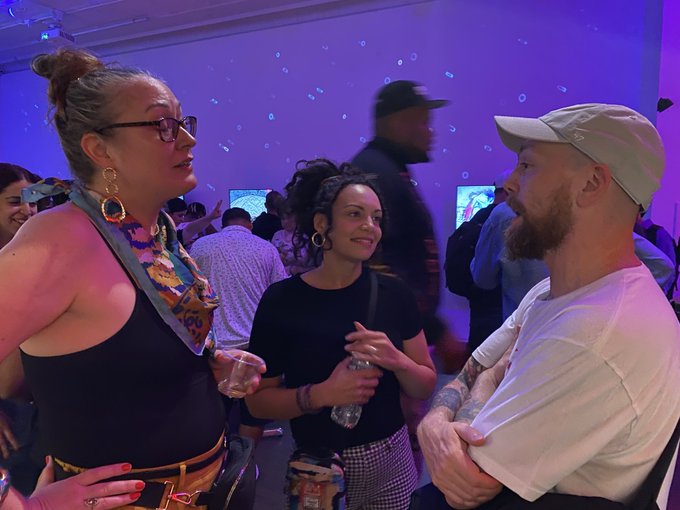 Three people converse in a room with purple lighting, including a woman with glasses, a smiling woman in a black shirt, and a bearded man wearing a cap.