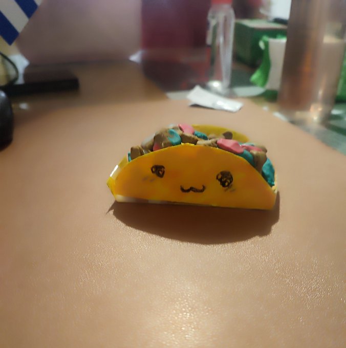 A small yellow taco-shaped object with a cute drawn-on smiling face and colorful filling (blue, pink, brown) sitting on a desk surface.