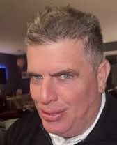 A close-up selfie of a man with short, styled blond hair wearing a dark jacket and light collared shirt, smirking confidently at the camera. His playful, self-assured expression fits the caption "Multiple performances you say?" suggesting a humorous nod to someone sporting different looks or styles.