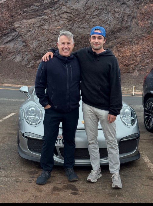 Two men pose together with their arms around each other in front of a silver Porsche sports car on a road with rocky cliffs in the background.
