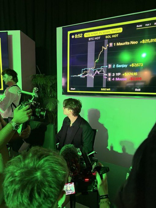 A scene at what appears to be a crypto or stock trading event with green lighting, showing a leaderboard screen displaying 'SOL.HOT' with rankings including '1 Maurits Neo +$25,81...' and '2 Sanjay +$7,673'.