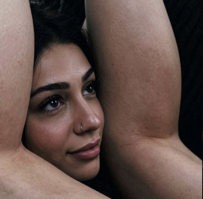 A close-up portrait of a woman with dark hair and a nose ring, her arms raised framing her face against a dark background.