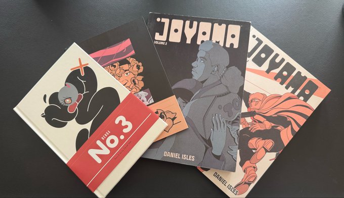 Several issues of the 'JOYAMA' comic book series by Daniel Isles are fanned out on a dark surface, including a Volume 2 issue and a Book No. 3.