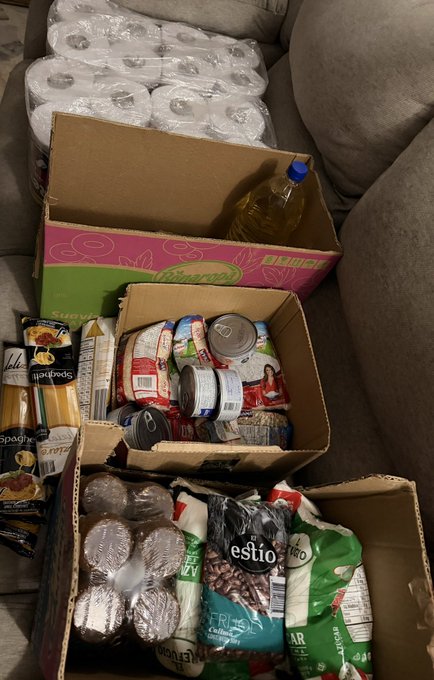 A collection of donated groceries sits on a couch, including bags of Nestlé Estilo coffee, packages of Bucatti spaghetti, canned goods, cooking oil in a bottle, and bulk packs of bottled water in a "Bucatti" branded box. The supplies, totaling around 220,000 Colombian Pesos (COP), reflect a community aid effort where contributors sent items to help supplement the recipient's $40 addition, showcasing tangible support through essential food and household products.