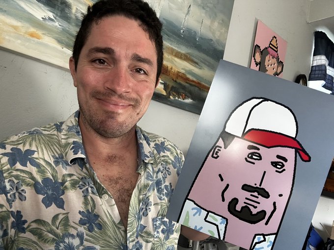A smiling man in a floral Hawaiian shirt holding a cartoon-style portrait painting of a man wearing a red and white cap.