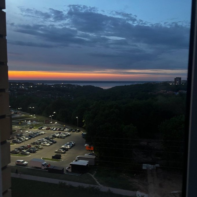 The image shows an aerial view of a parking lot filled with cars at dusk. The sky above has a beautiful sunset color, creating a serene atmosphere over the scene. There are several cars parked in various positions within the lot, indicating that this is likely a popular location for people to park their vehicles while enjoying the view or attending nearby events.