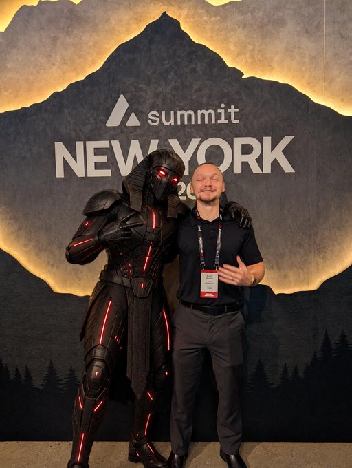 A man in a black polo shirt with a lanyard poses next to a person in an elaborate dark futuristic samurai-style armor costume with glowing red accents, standing in front of a 'summit NEW YORK' backdrop featuring a mountain logo.