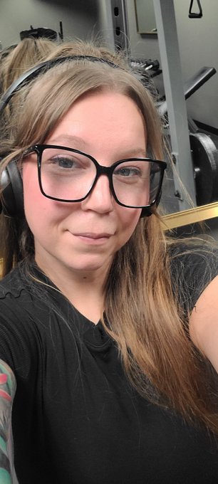 A selfie of a person with glasses and headphones wearing a black shirt, taken inside what appears to be a gym with equipment visible behind them.