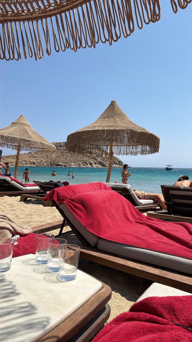 A sunny beach scene featuring straw umbrellas, lounge chairs with red towels, turquoise water with swimmers, and a table in the foreground holding two glasses of water.
