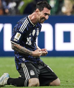 Lionel Messi in an Argentina national team jersey kneeling on a soccer field with an emotional expression.
