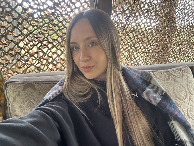 A young woman with long, highlighted blonde hair poses for a selfie in a cozy outdoor lounge area, wrapped in a dark sweatshirt and tartan plaid blanket, with a decorative camouflage netting backdrop. The image complements a cozy evening greeting, suggesting a peaceful night spent relaxing in nature.