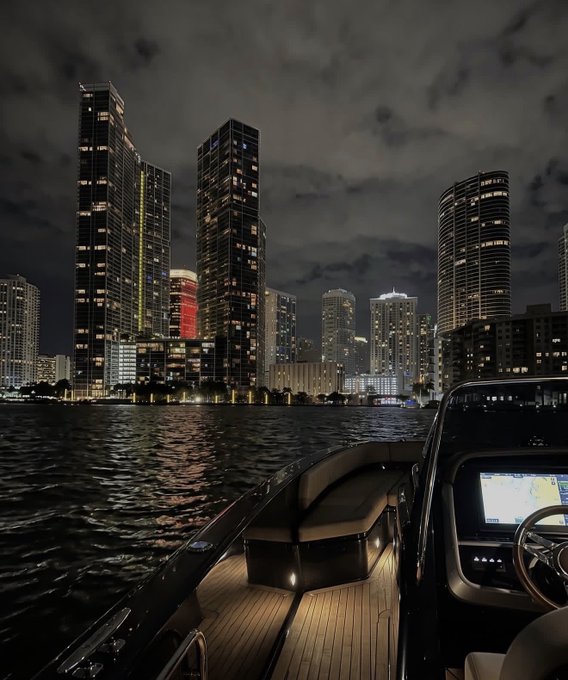 A nighttime view from a luxury boat shows a wooden deck and illuminated control panel, with a city skyline of glowing skyscrapers across the water under a cloudy sky.