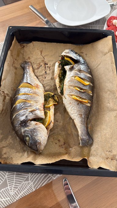 Two whole baked fish with lemon slices stuffed inside, served on parchment paper in a black baking tray on a wooden dining table.