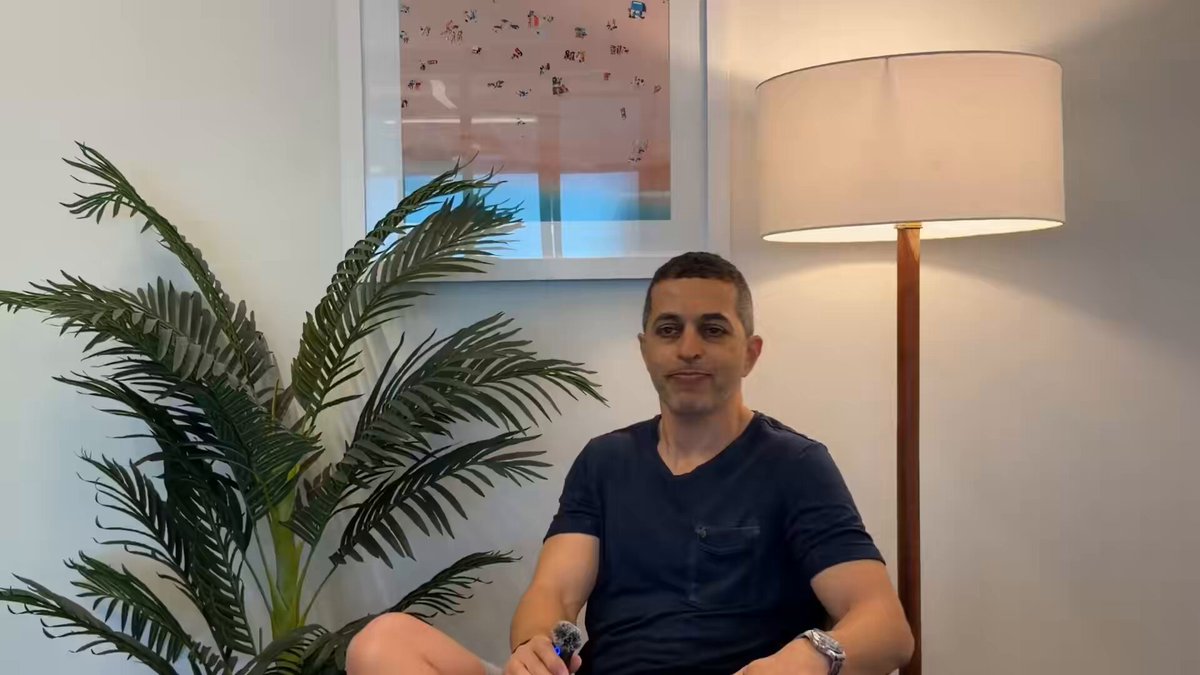 The image features a man sitting down with his legs crossed while wearing a blue shirt. He has a pen in his hand, possibly taking notes or writing something down. The room also contains a lamp that illuminates the scene, creating a cozy atmosphere. In the background, there is a picture hanging on the wall and another plant placed nearby. The image includes text that reads "vibecoding, ai layoffs, where eth lands in 2 years @GuyItz doesn't dodge any of it in this video".