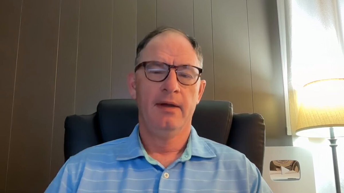 The image shows a man wearing glasses sitting in an office chair, with his head resting on the armrest of the chair. The background features a wall with vertical blinds that allow natural light into the room. There are also two lamps present in the scene - one located near the center and another towards the right side of the frame.