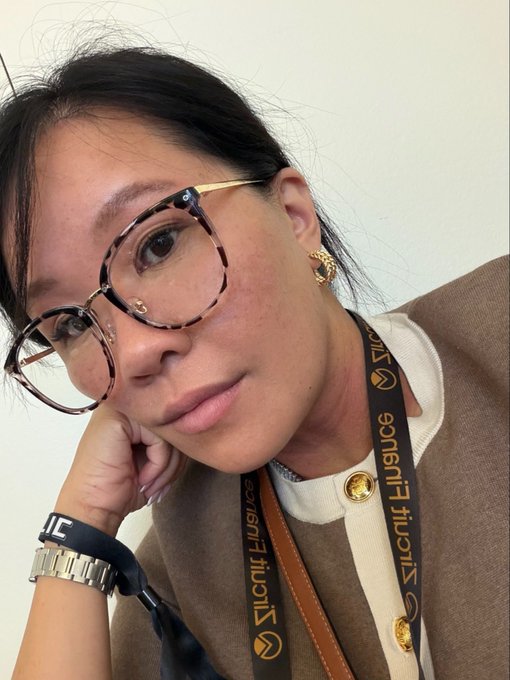 A selfie of a woman wearing tortoiseshell glasses and a beige blazer with gold buttons, with a 'VICTORIA LIANG' lanyard around her neck.