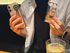 A person taking a mirror selfie while holding a phone and a small decorative figurine, with a glass of beverage in the foreground.