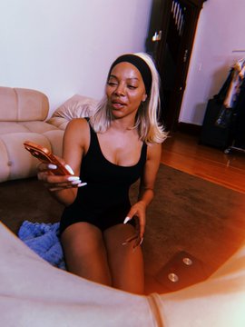 A woman with blonde hair in a black bodysuit and white headband holds a smartphone while sitting on a cream-colored couch.