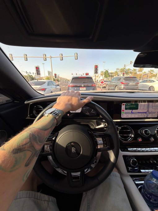 A tattooed arm grips the steering wheel of a Rolls-Royce at a traffic light, with the dashboard navigation displaying directions, contrasting the post's playful threat about "liquidating" GM.