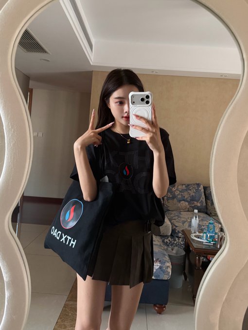 A young woman takes a mirror selfie wearing a black t-shirt and pleated skirt, holding a phone and making a peace sign, with a tote bag reading 'HXL DAO' visible.