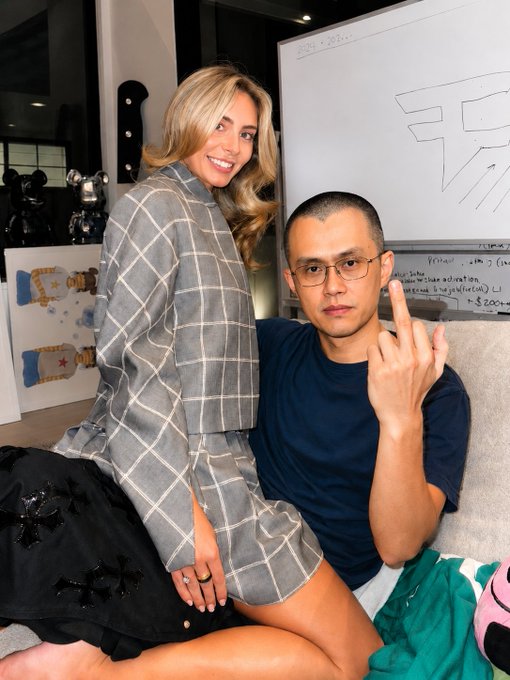 The image features a man sitting on a couch with his arm raised, making a peace sign gesture while wearing glasses and a blue shirt. A woman is also present in the scene, sitting next to him. They are both smiling for the camera. The background of the photo includes a whiteboard that has some writing on it, as well as a few other people standing around them. There's an additional person standing behind the couch who appears to be wearing a black and white shirt with a design on it.