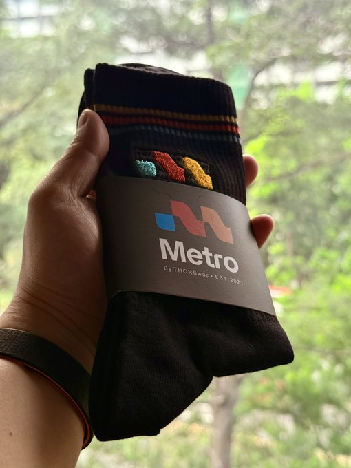 A hand holds a pair of black "Metro" branded socks by THORSwap (Est. 2021) up against a window with a blurred green forest view, likely captured as a swag giveaway at Token2049 in Singapore to promote Onchain conversations.