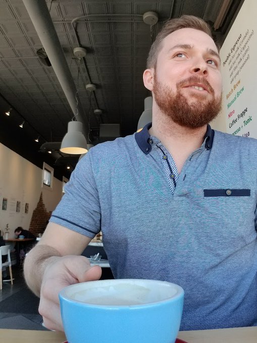 A bearded man in a blue polo shirt sits in a café, looking up with a slight smile as he holds a latte in a teal cup, with a handwritten menu board in the background listing items like "Buttermilk Pancake," "PB&Brew," and "Coffee Frappe." The casual, relaxed moment paired with the caption's playful "GM future millionaires" gives the photo a cheerful, aspirational vibe, as if he's sipping a morning coffee while dreaming about future success.