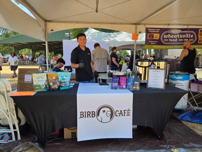 A photo of a young man standing behind a table with a 'BIRB CAFÉ' banner at an outdoor event, giving a thumbs up, with products displayed and a 'wheatsville food co-op' booth visible in the background.