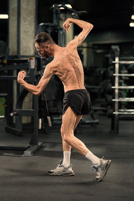 A shirtless, lean man posing in a gym wearing black shorts, white socks, and athletic shoes, displaying visible muscle definition.